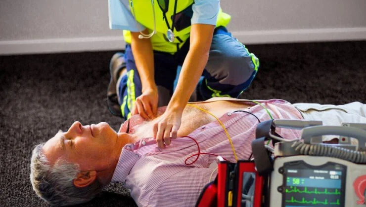 La imagen muestra una situación de emergencia real. Se ve a un paramédico arrodillado junto a un hombre mayor que está acostado en el suelo. El paramédico parece estar examinando al hombre, quien tiene cables conectados a su pecho, posiblemente de un monitor cardíaco o un desfibrilador. El paramédico lleva un chaleco de alta visibilidad.