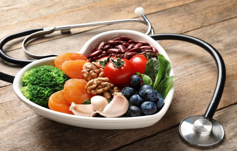 Un plato en forma de corazón lleno de alimentos saludables como frutas, verduras, nueces y frijoles, junto a un estetoscopio. Representa una alimentación saludable para el corazón y por ende la salud cardiovascular.