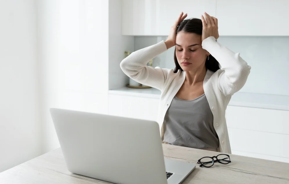 Una mujer joven frente a una computadora portátil se lleva las manos a la cabeza. Su expresión y postura sugieren estrés o agotamiento. Esta imagen ilustra el impacto del estrés digital en el bienestar, un factor importante para la salud cardiovascular.