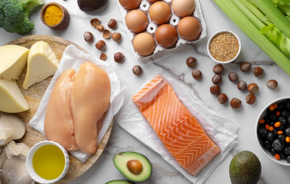 Una variedad de alimentos ricos en diferentes tipos de grasas están dispuestos sobre una superficie de mármol. Se observan pechugas de pollo crudas, un filete de salmón, aguacates, huevos, queso, aceite de oliva, nueces, semillas de lino y arándanos. La diversidad de estos alimentos ilustra la idea de que existen diferentes tipos de grasas, algunas beneficiosas y otras que deben consumirse con moderación.