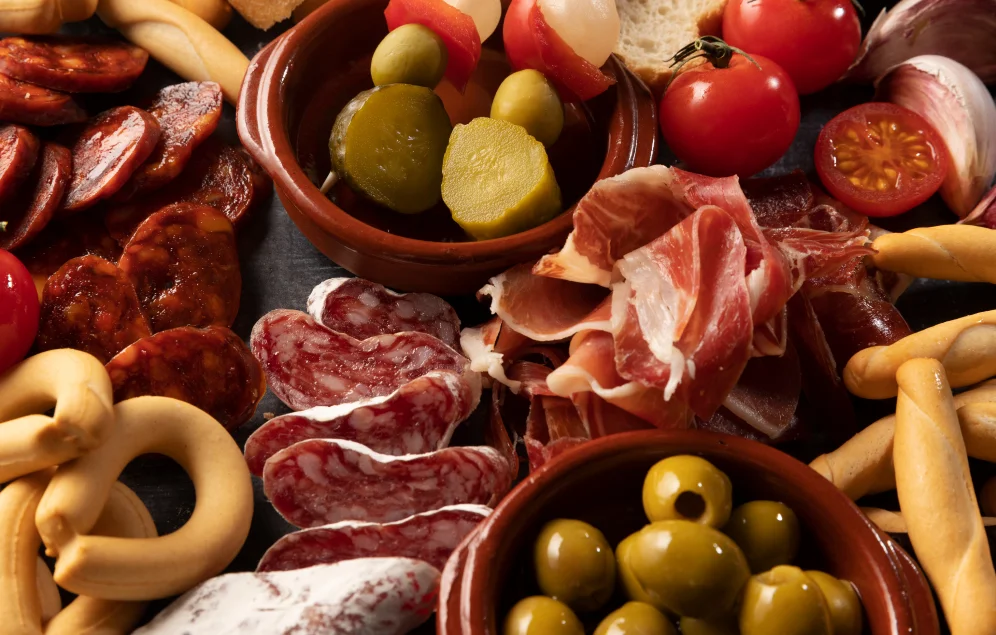 Una selección de alimentos procesados y ricos en grasas saturadas y sodio se presenta en varios recipientes sobre una superficie oscura. Se observan rodajas de chorizo, salchichón, jamón curado, aceitunas, pepinillos en vinagre, tomates cherry, ajo y pan tipo colines. Este tipo de alimentos, consumidos en exceso, podrían ser perjudiciales para la salud cardiovascular y, por lo tanto, deberían evitarse o consumirse con moderación si existe preocupación por el corazón.
