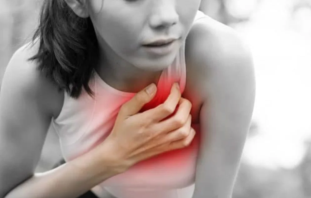 Una mujer joven parece experimentar dolor o malestar en el pecho, ya que se lleva la mano al centro del tórax. El área donde se encuentra su mano está resaltada en rojo, lo que visualmente enfatiza la posible ubicación del dolor. Su expresión facial sugiere incomodidad o preocupación. Esta imagen podría ilustrar síntomas relacionados con problemas cardíacos