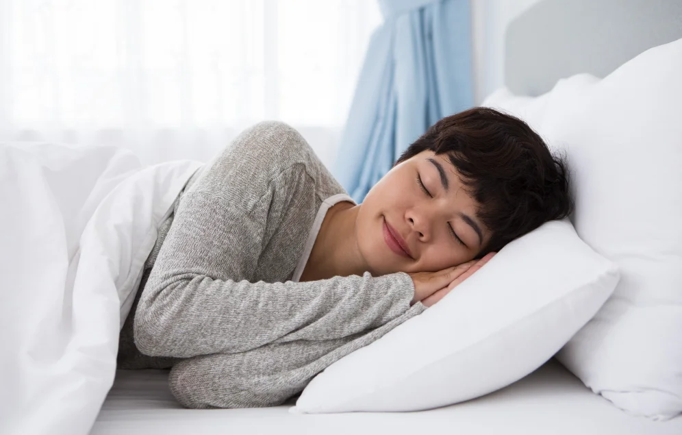 Una persona de apariencia joven, con cabello corto y oscuro, duerme plácidamente de lado, apoyada en una almohada blanca y cubierta con una sábana blanca. Su rostro relajado y sus ojos cerrados sugieren un sueño profundo y reparador. La escena evoca la importancia del descanso adecuado para el bienestar general, incluyendo la protección de la salud cardiovascular.